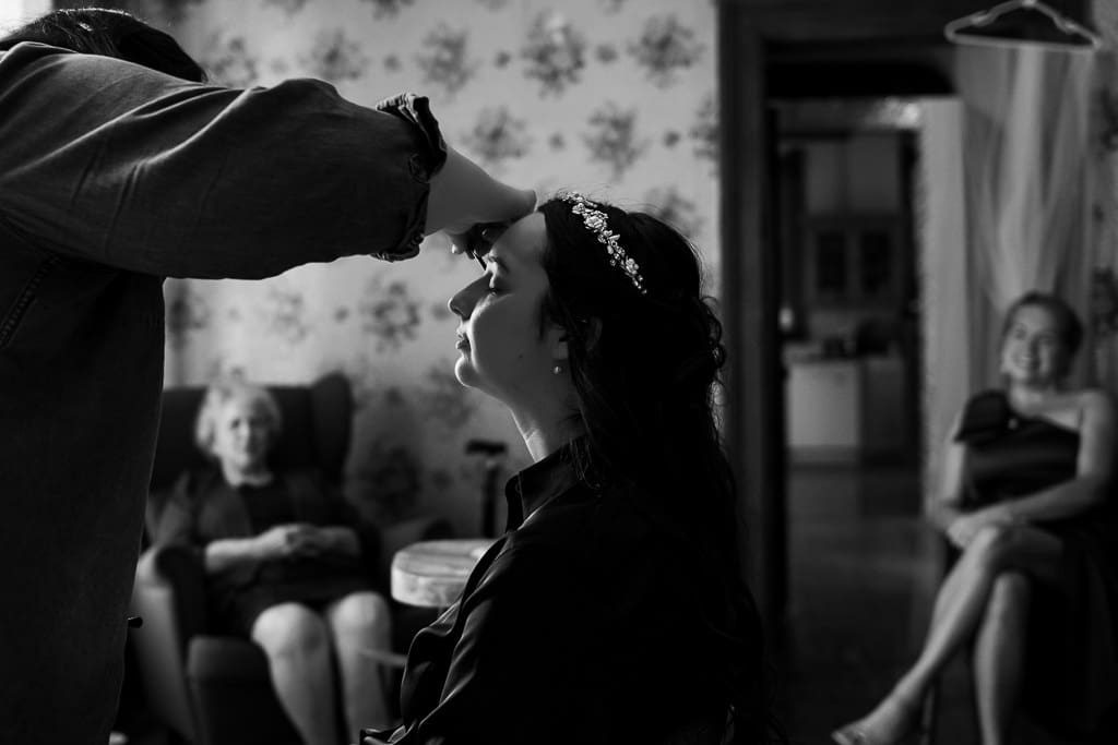 Bride getting her makeup done before her wedding. Family watching in the background.