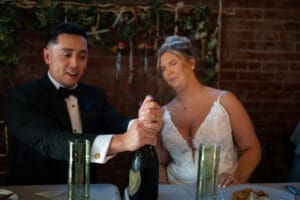 Groom popping champagne while the bride reacts with surprise, closing her eyes