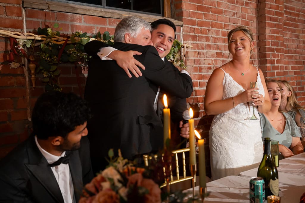 Groom hugging his dad after his speech, while the bride stands next to them laughing during a multicultural wedding at Union Kansas City