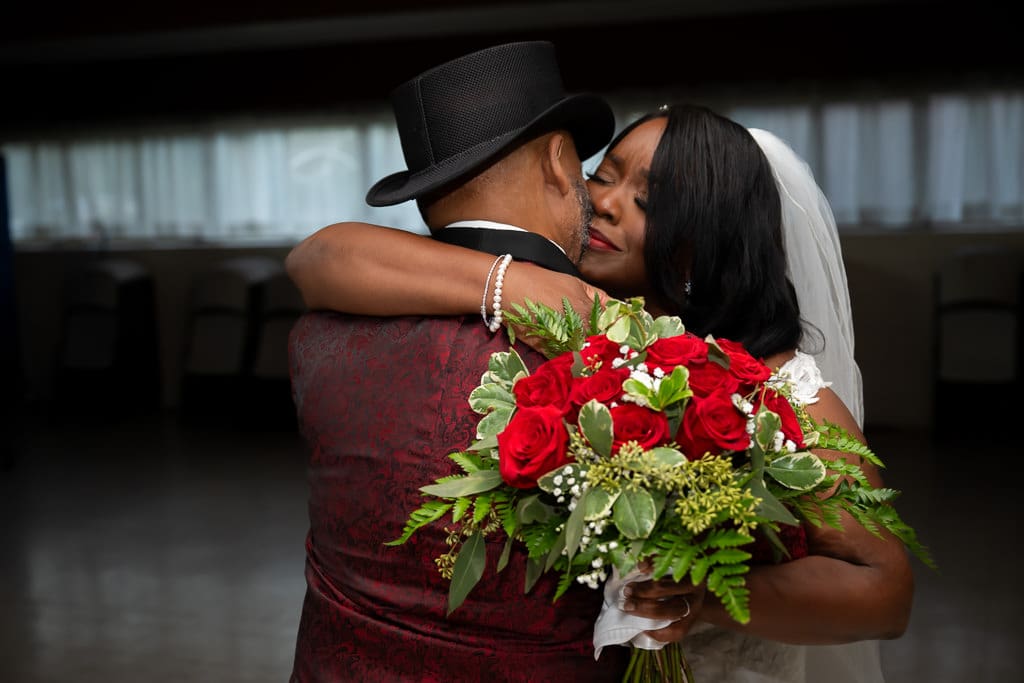 Leona hugs Michael tightly after the ceremony, red rose bouquet in hand, his black top hat against her veil