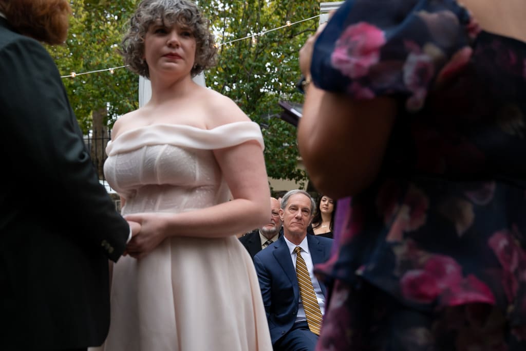 Father in focus in the background with emotion while the bride looking toward the officiant during the wedding ceremony blurry in the foreground