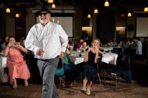 Father of the bride in a black hat and sunglasses strutting past laughing guests at a Boulevard Brewing Company wedding