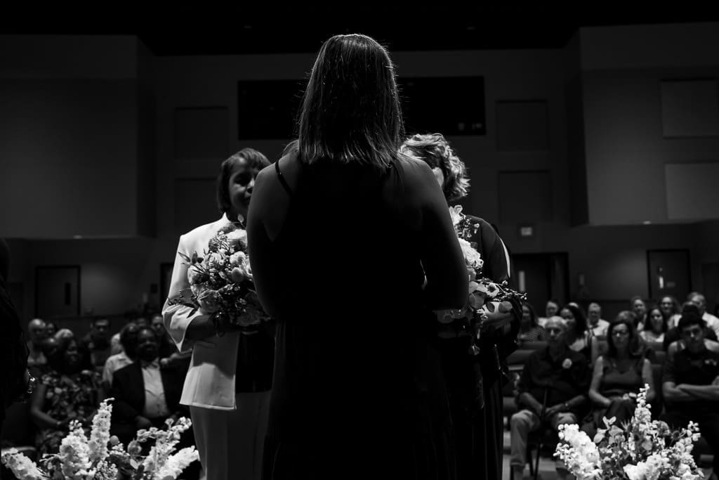 Black and white image of the officiant in the foreground with the two brides partially hidden during their wedding ceremony