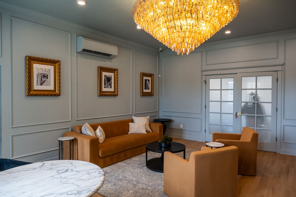 Lounge at Melrose Abbey with a gold chandelier, framed photos and caramel velvet chairs