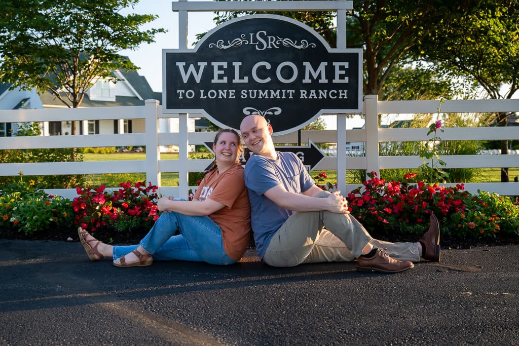 Elsie and Jason sit under the Welcome to Lone Summit Ranch sign, red flowers behind them