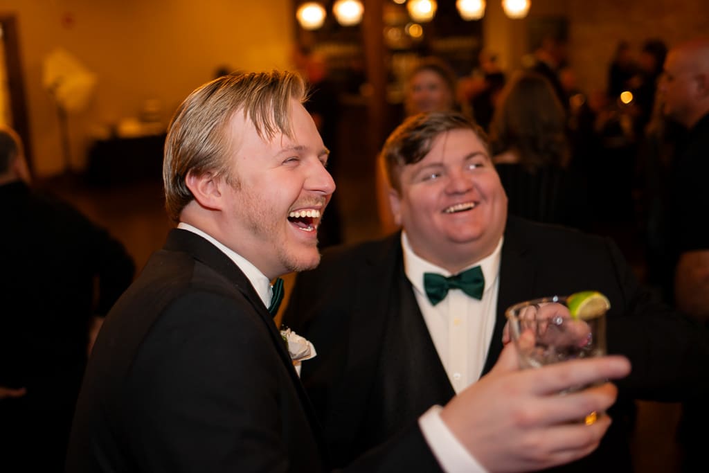 Two groomsmen laugh and raise their drinks during the reception at The Stanley