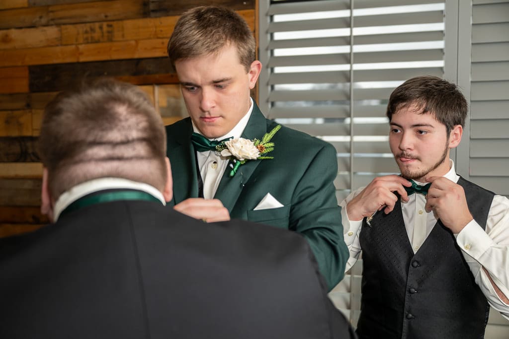 Drake helps a groomsman with his bow tie while another groomsman fixes his own, tongue sticking out